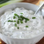 Creamy cucumber dip with fresh chives in a white bowl, served with crackers and a spoon.