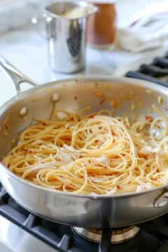 Cooked pasta and shredded chicken being combined in a stainless steel skillet with red pepper flakes.
