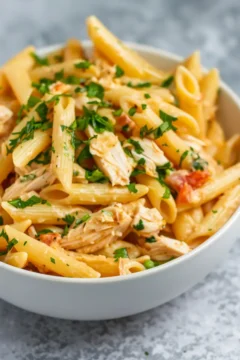 Creamy penne pasta with shredded rotisserie chicken and herbs in a white bowl.