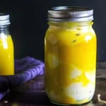 Jar of mustard pickled eggs in a vibrant yellow brine, placed on a wooden surface with a second jar in the background.