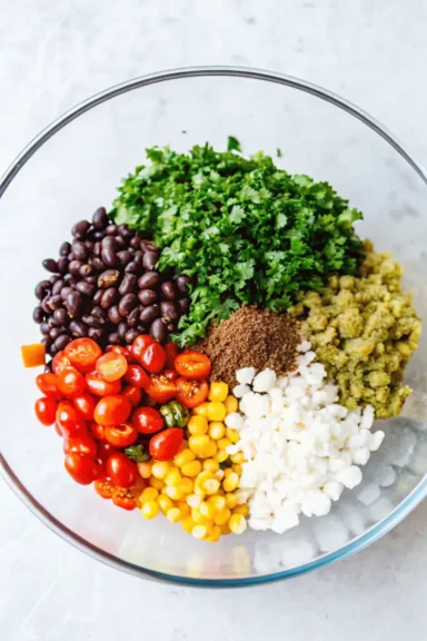 A glass bowl filled with fresh ingredients for Redneck Caviar, including black beans, cherry tomatoes, corn, cilantro, and spices.