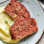 Close-up of two slices of juicy meatloaf with a glossy ketchup glaze, served over creamy mashed potatoes.