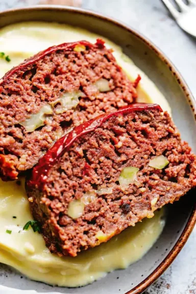Close-up of two slices of juicy meatloaf with a glossy ketchup glaze, served over creamy mashed potatoes.