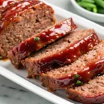 Sliced BBQ meatloaf outrageously flavorful with a rich, glossy glaze, served on a white platter with fresh herbs.