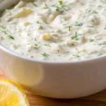 Creamy homemade tartar sauce with fresh dill and lemon in a white bowl on a wooden board.